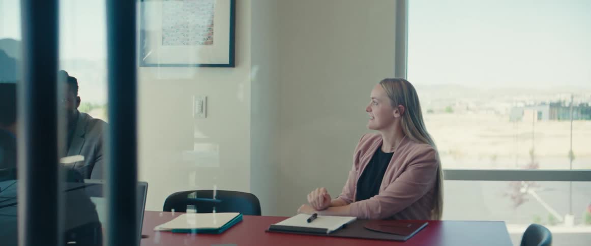 A business meeting in a glass office with a man standing and pacing and the other people sitting at a table