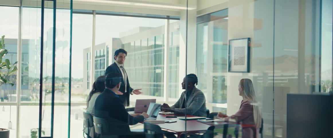 A man talks at an office meeting with four other people sitting at a table with open laptops