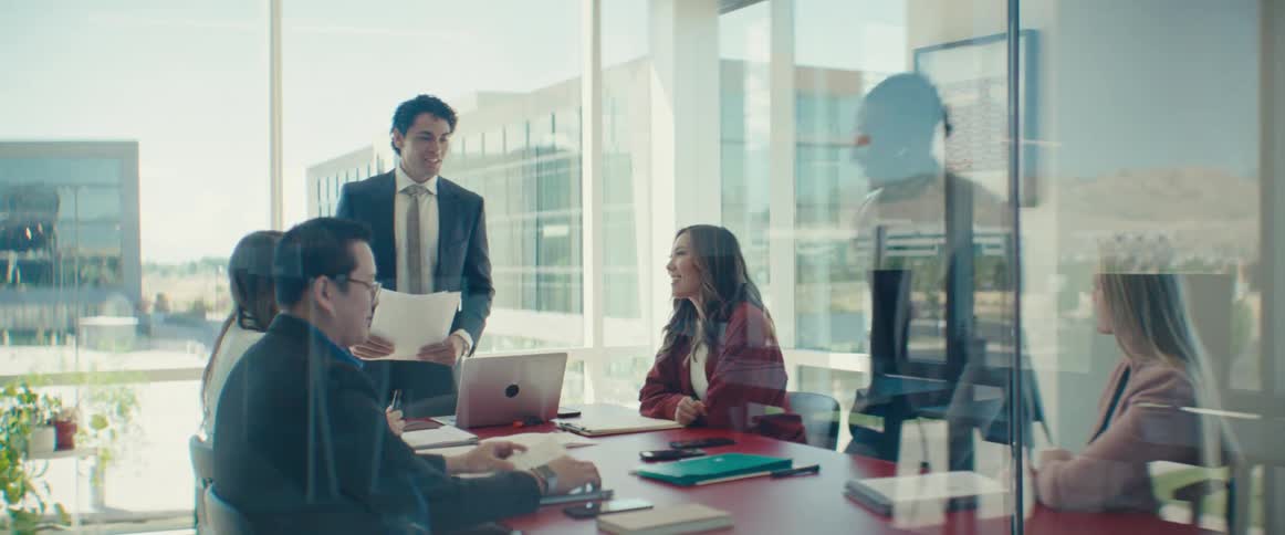 Man sitting at a table in an office meeting with another man standing talking and holding a document in his hands