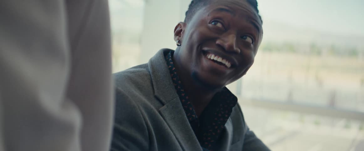 A black man smiles while talking to another person in an office setting