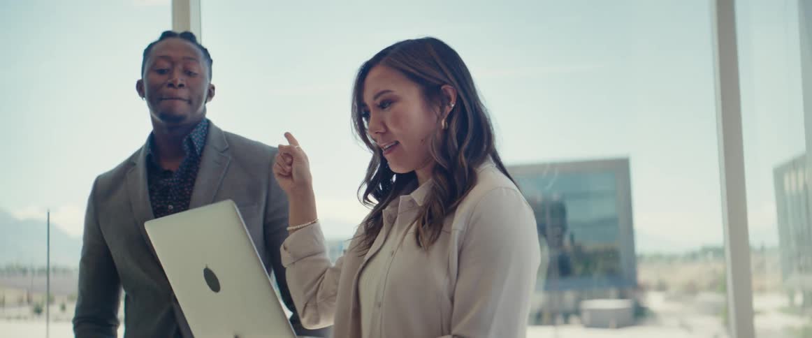A woman passionately explains a point to a man while they both look at a laptop in a modern office setting with a city view
