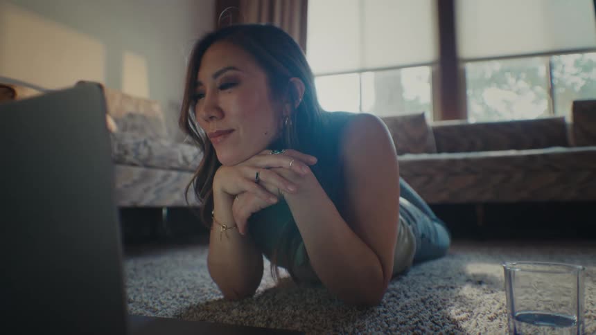 Woman lying on the living room rug and interacting with a laptop in front of her