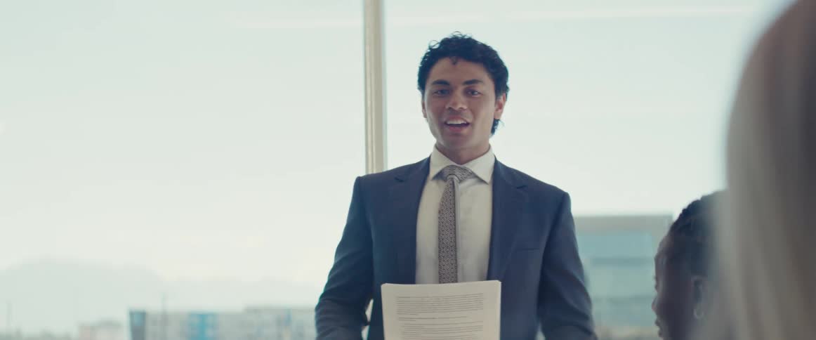 Man in a suit standing holding a document talking to a female colleague who is sitting at a table