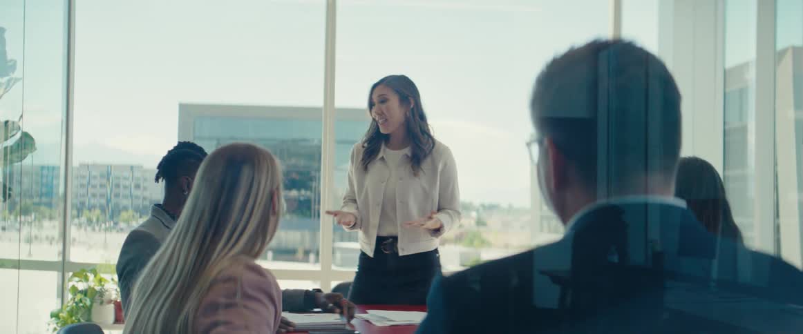 A woman stands and presents to a group of people in a modern office setting with large windows