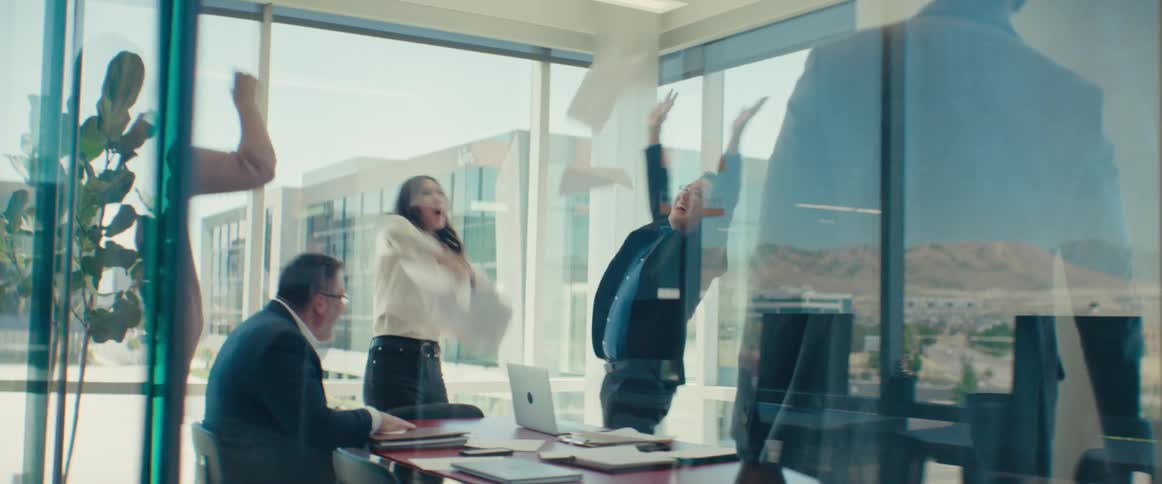 A business meeting in a glass office with everyone throwing their papers in the air and wooting in celebration