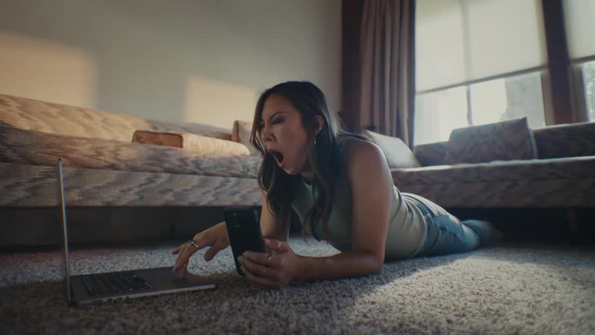 A woman is laying on the floor looking at her cell phone and laptop