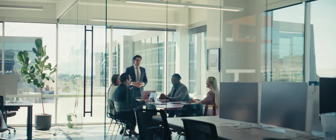 A group of business professionals are gathered around a table in a modern office with a man leading a presentation