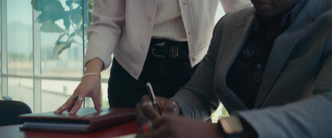 Man sitting at an office desk writing and a woman standing next to him interacting