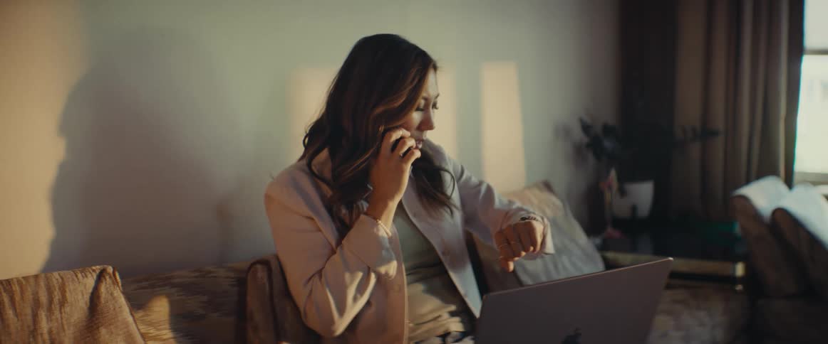 A woman sitting on her couch talking on her cell phone while looking at her laptop before getting off the couch
