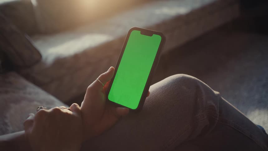 An adult sits indoors on a couch while scrolling on a smartphone