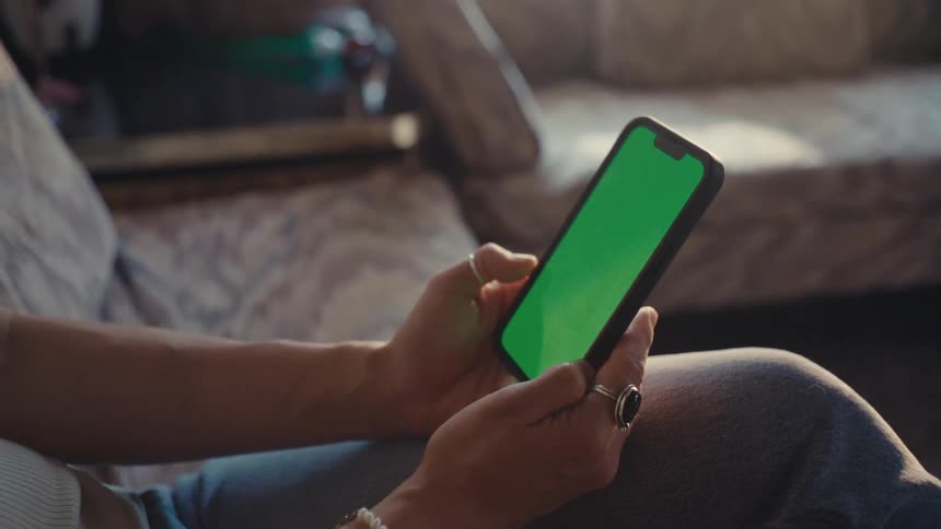 A person holds a smartphone with a green screen in an indoor setting with warm lighting