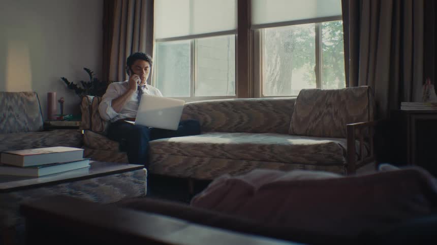 Man sitting on the couch picking up a smartphone and answering a phone call while interacting with a laptop