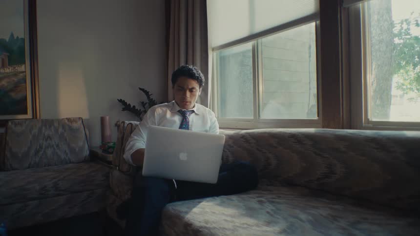 A man sitting on a couch wearing a shirt and a tie with a laptop on his lap looking worried