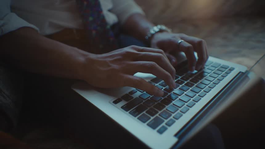 Close up of a man typing on a laptop keyboard as he sits on a couch