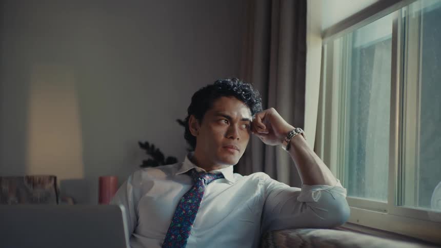 A man sitting on a couch by a window wearing a shirt and tie and with a laptop on his lap gazing out of the window in contemplation