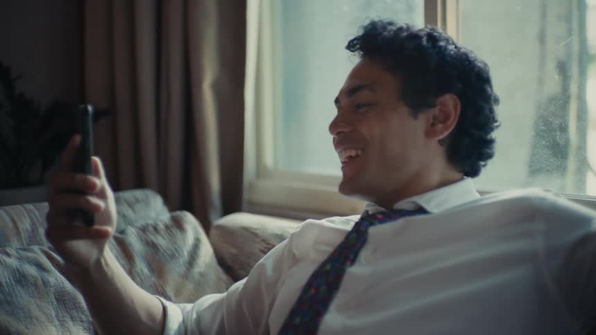 A young man sitting on a couch holding his cellphone in front of him having a conversation with someone on the other end