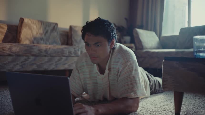 A man lying on the floor of a living room works on a laptop and looks worried until he has a thought and smiles