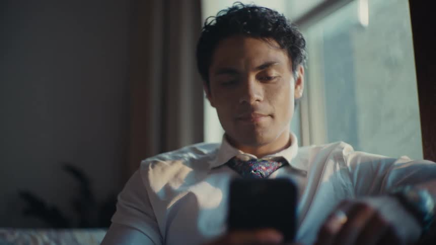 A young man in a white shirt and tie is looking at a cell phone in his hand and he is sitting