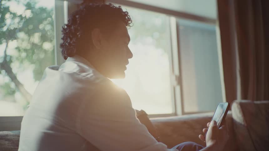 An young man with curly hair is sitting by a window holding a smartphone having a video call