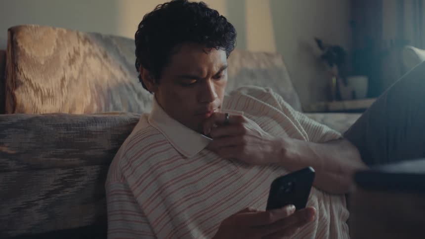 A man lounging in his living room while using his cellphone and looking at itcontemplatively