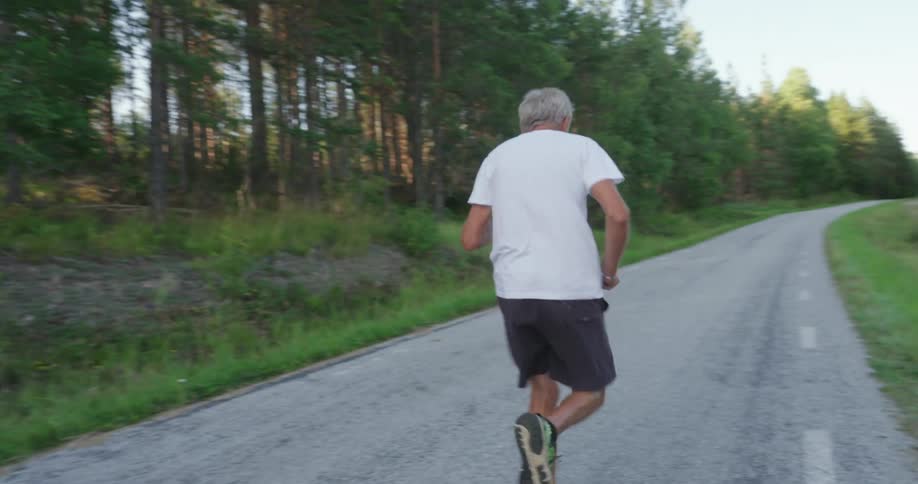 An older man running on a roadway as the sun sets behind the trees lining the road