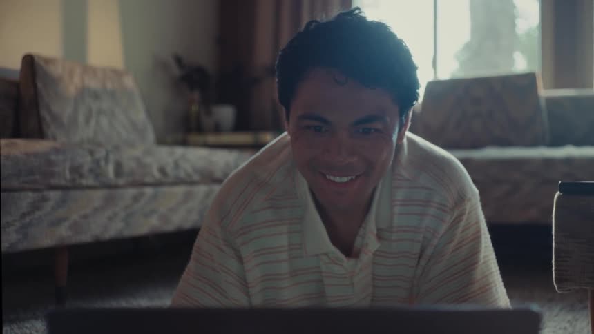 A man with a happy expression is seen sitting on the floor looking at a laptop in a living room setting
