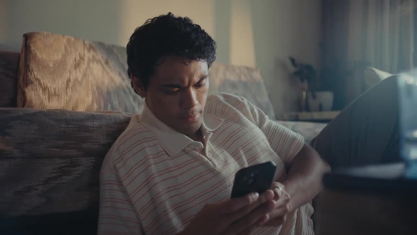 A man sitting on the floor with his back leaning against a couch looking worried as he holds a smart phone