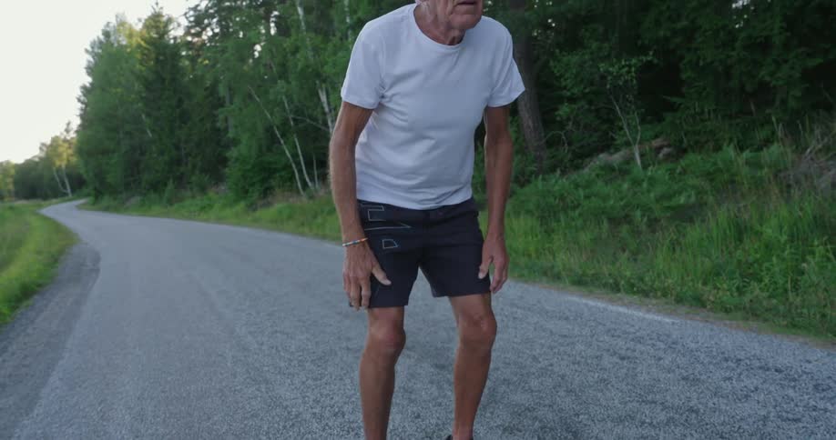 An elderly man stops on a path near a forest to catch his breath after jogging