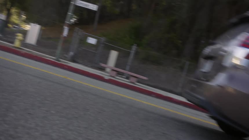 A gray car driving on the road near a sidewalk and fence along a tree line