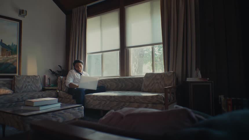 A man sits on a couch with his laptop on his lap talking to someone