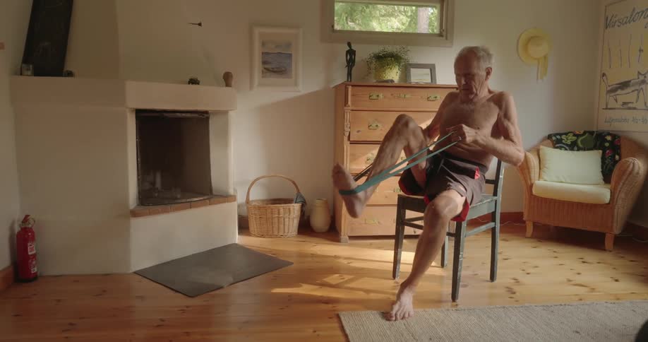 An elderly man sits on a chair and uses exercise bands with his legs