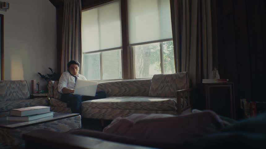 A young man sitting on a couch at home working on a laptop while talking on the phone