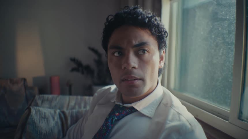 A man sitting on a couch with his back to a window wearing a shirt and a tie looking worried