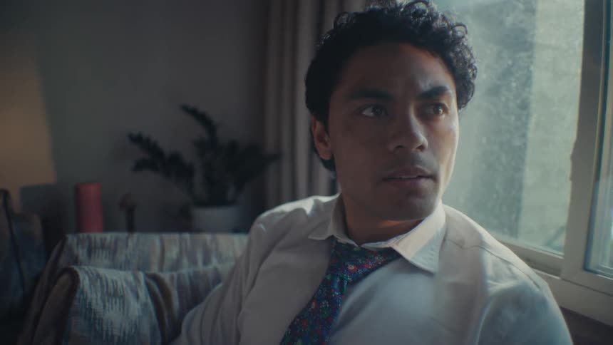 Close up of a man sitting on a couch next to a window looking contemplatively wearing a dress shirt and tie