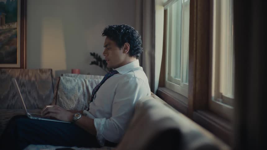 A man sits on a couch in a room typing on a laptop with a window behind him