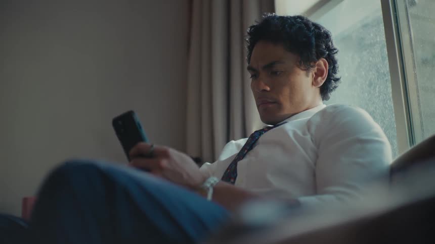 A man sitting on his couch in front of a window while looking at his phone before looking around the room