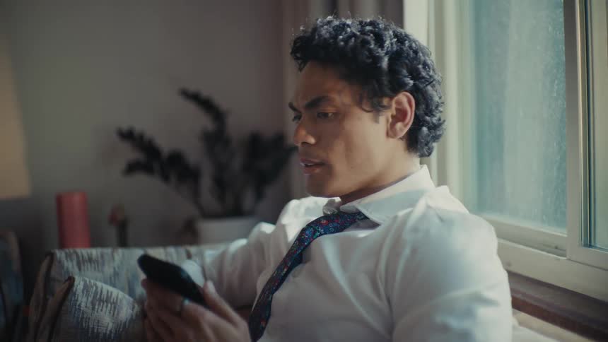 An young man with curly hair is sitting by a window holding a smartphone having a call