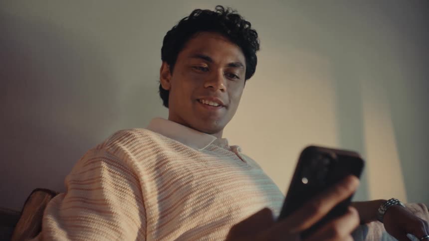 A young man sitting on a couch looking at his cellphone shaking his head and smiling