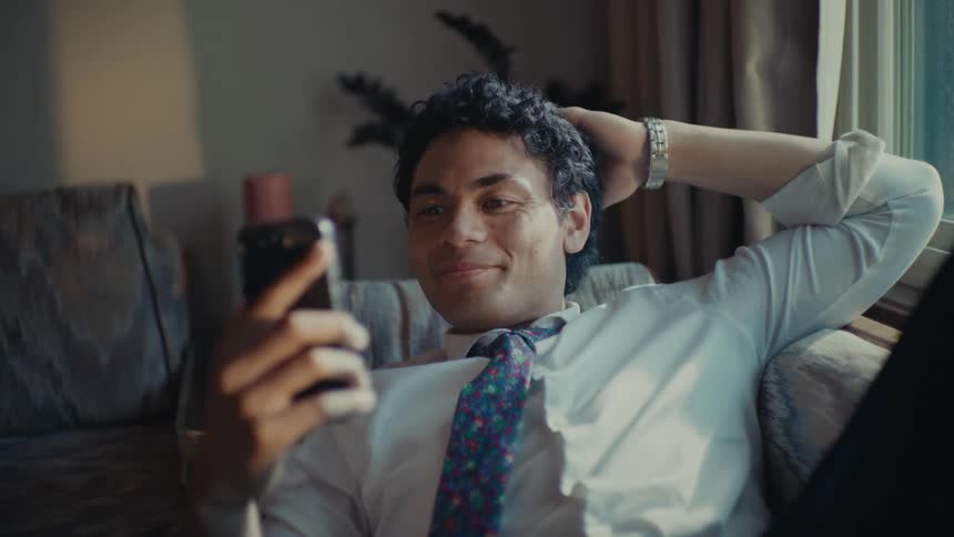 A man in formal attire rests on a couch at home talking on his phone