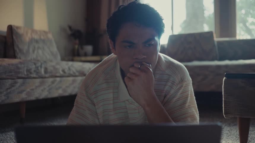 A young man laying on the living room floor in front of a laptop while thinking and looking off to the right
