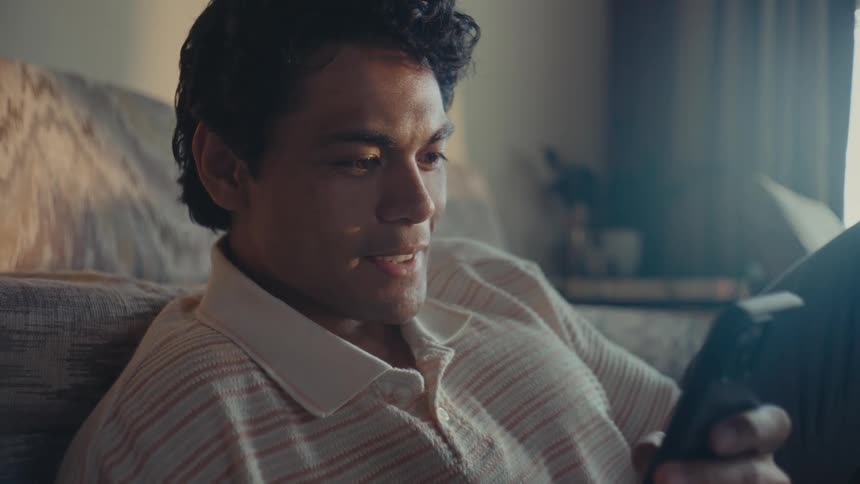 A man sitting on the floor of a living room leaning against a couch smiles as he reads from his smart phone