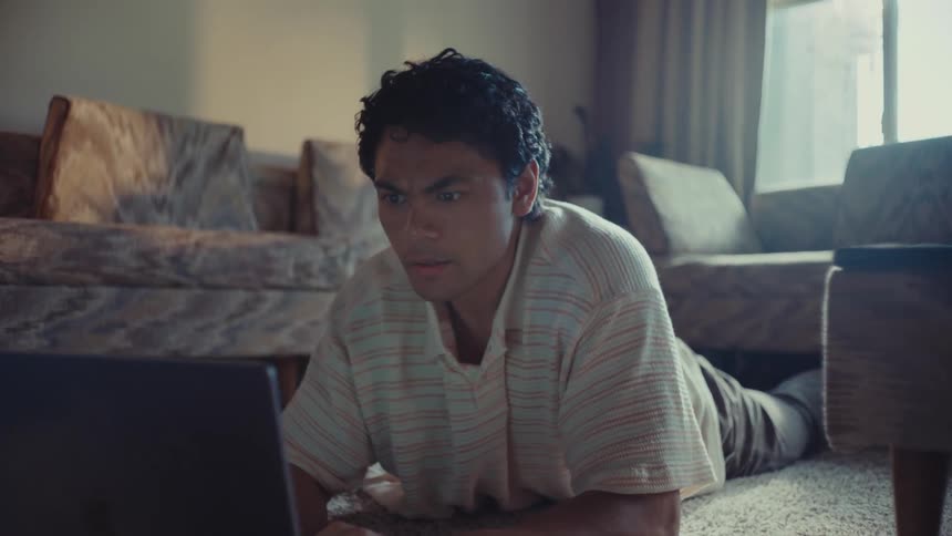 A young man is lying on the carpet in a living room and is using a laptop and has a surprised expression