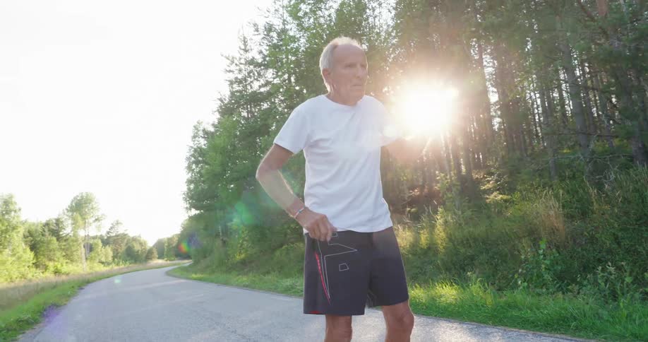 An elderly man in a white shirt is standing on a rural gravel road and he is stretching his arm