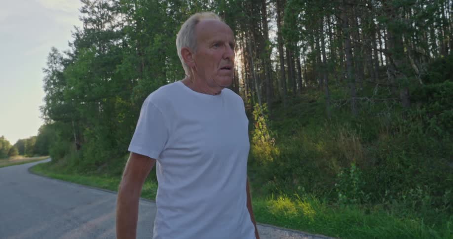 An elderly man in a white shirt stands on a concrete street with vegetation on either side
