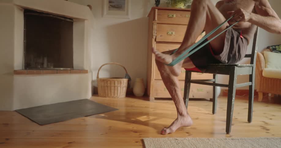 Shirtless elderly man sitting on a chair and using an elastic band to stretch his leg