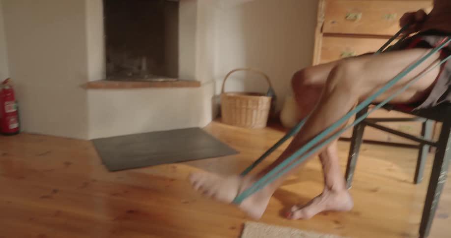 An elderly man sits on a chair while using exercise bands with his legs