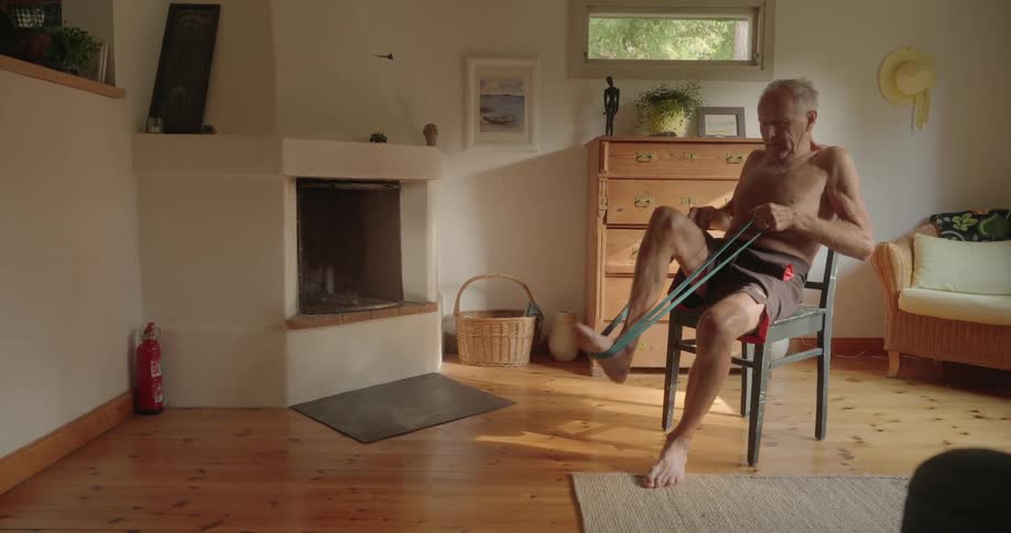 Elderly man sitting on a chair in the living room using a rubber band and stretching his legs