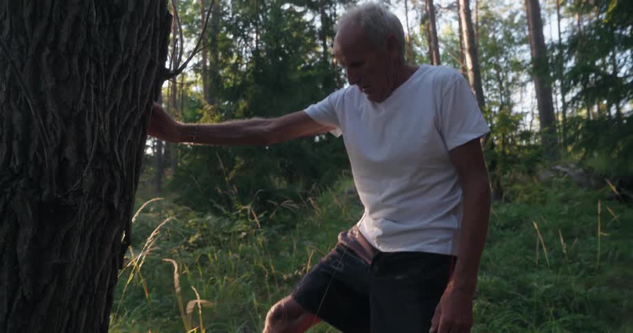 An elderly man doing leg warmup exercises while leaning against a tree
