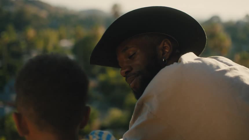 A father talks to his little boy outside while wearing a cowboy hat and sitting down
