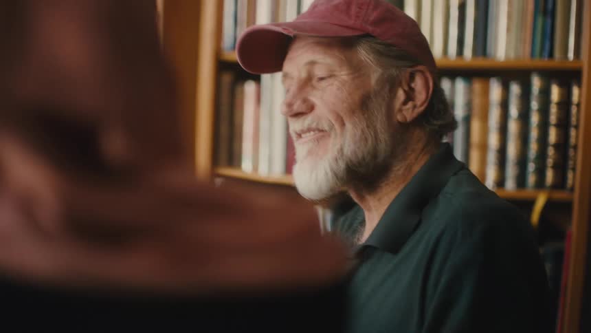 Elderly man talking to another person while sitting in a library with a book in the background